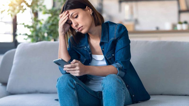 Shot of worried young woman using her mobile phone while thinking about problems sitting on couch in the living room at home.