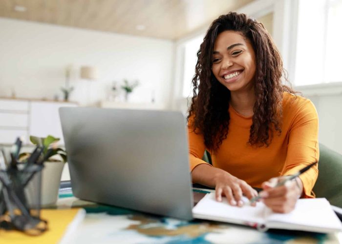 Distance Education. Portrait of smiling woman sitting at desk, using laptop and writing in notebook, taking notes, watching tutorial, lecture or webinar, studying online at home looking at screen